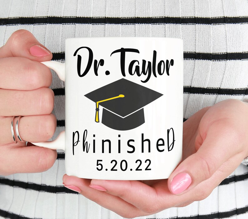 May include: White ceramic mug with black text that reads "Dr. Taylor Phinished 5.20.22". A black graduation cap with a gold tassel is above the text.