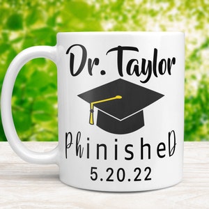 May include: White ceramic coffee mug with a black graduation cap and the text "Dr. Taylor Phinished 5.20.22"