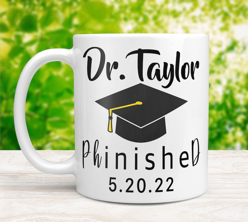 May include: White ceramic coffee mug with a black graduation cap and the text "Dr. Taylor Phinished 5.20.22"