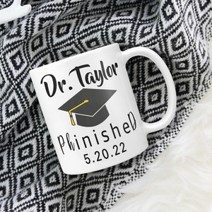 May include: White ceramic mug with black text that reads "Dr. Taylor finished 5.20.22" and a black graduation cap with a yellow tassel.