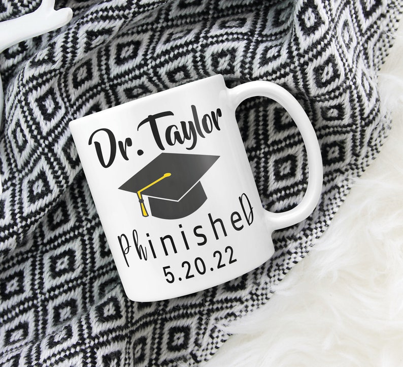 May include: White ceramic mug with black text that reads "Dr. Taylor finished 5.20.22" and a black graduation cap with a yellow tassel.
