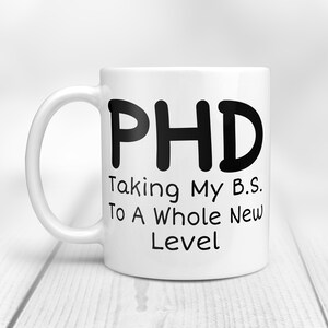 Puede incluir: Taza de caf&eacute; de cer&aacute;mica blanca con texto negro que dice "PHD Taking My B.S. To A Whole New Level".