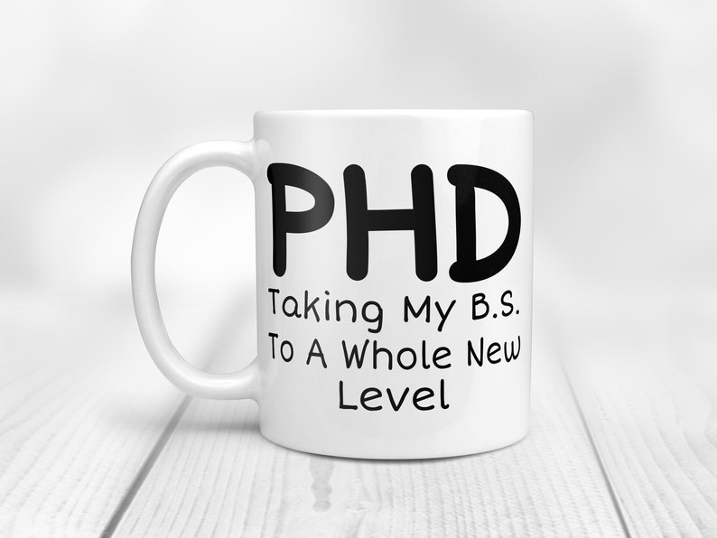 Puede incluir: Taza de caf&eacute; de cer&aacute;mica blanca con texto negro que dice "PHD Taking My B.S. To A Whole New Level".