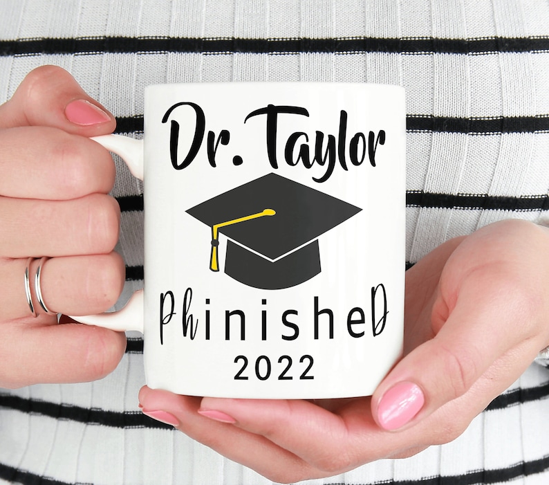 May include: White ceramic mug with black text that reads "Dr. Taylor Phinished 2022" and a black graduation cap with a gold tassel.
