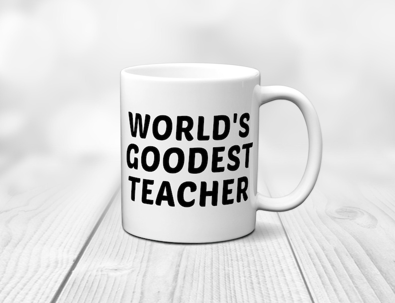 May include: White ceramic mug with the text "WORLD'S GOODEST TEACHER" in bold black letters. The mug has a curved handle and is set against a blurred background on a light wooden surface.