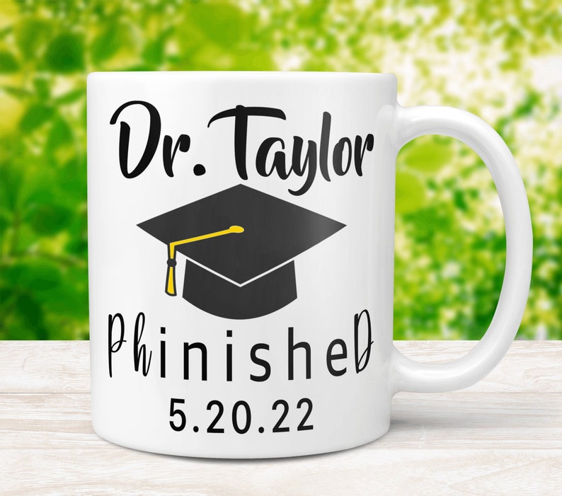 May include: White ceramic mug with black text that reads "Dr. Taylor Phinished 5.20.22" and a black graduation cap with a yellow tassel.