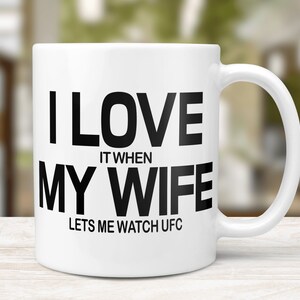 Pode incluir: Caneca de caf&eacute; de cer&acirc;mica branca com um gr&aacute;fico preto que diz "Eu amo quando minha esposa me deixa assistir UFC".