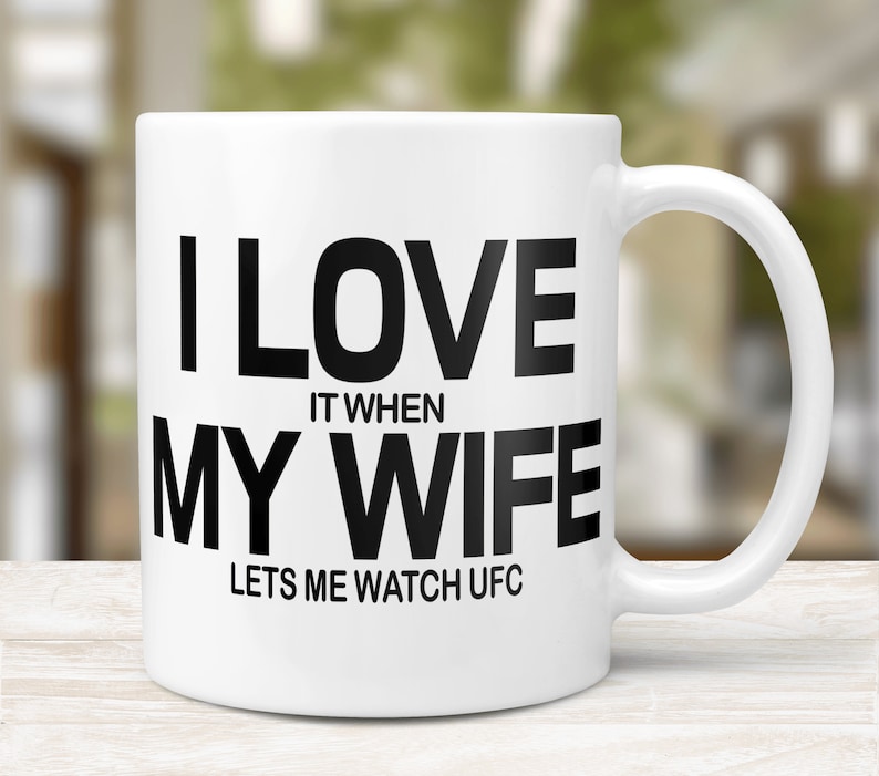 Pode incluir: Caneca de caf&eacute; de cer&acirc;mica branca com um gr&aacute;fico preto que diz "Eu amo quando minha esposa me deixa assistir UFC".