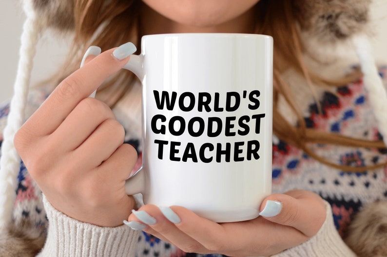 May include: A white ceramic mug with the text "WORLD'S GOODEST TEACHER" in bold black letters. The mug is held by a person wearing a patterned sweater and a white hooded jacket.