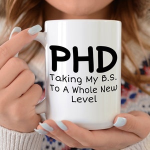 Puede incluir: Taza de caf&eacute; de cer&aacute;mica blanca con texto negro que dice "PHD Taking My B.S. To A Whole New Level"