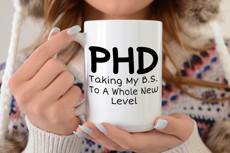 Puede incluir: Taza de caf&eacute; de cer&aacute;mica blanca con texto negro que dice "PHD Taking My B.S. To A Whole New Level"
