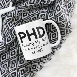Puede incluir: Taza de caf&eacute; de cer&aacute;mica blanca con texto negro que dice "PHD Taking My B.S. To A Whole New Level". La taza est&aacute; sobre una manta de patr&oacute;n blanco y negro.