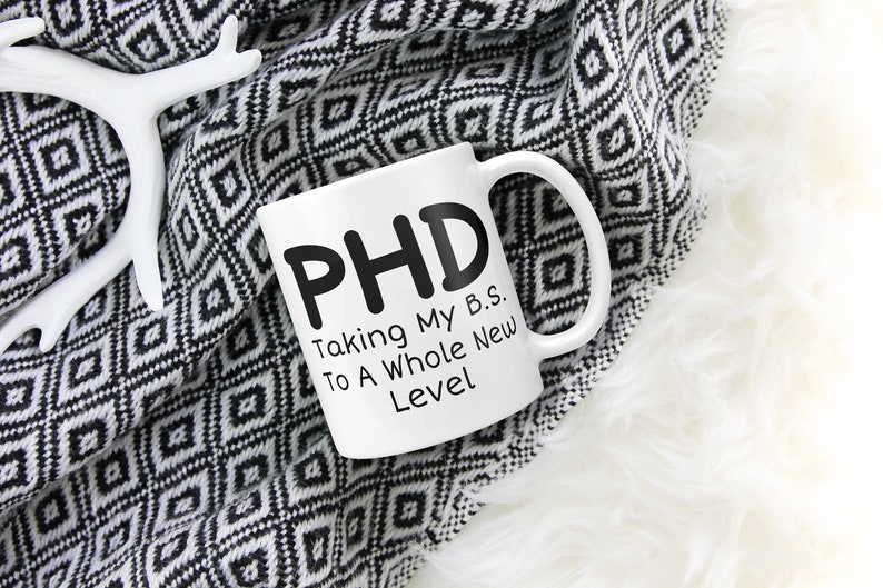 Puede incluir: Taza de caf&eacute; de cer&aacute;mica blanca con texto negro que dice "PHD Taking My B.S. To A Whole New Level". La taza est&aacute; sobre una manta de patr&oacute;n blanco y negro.