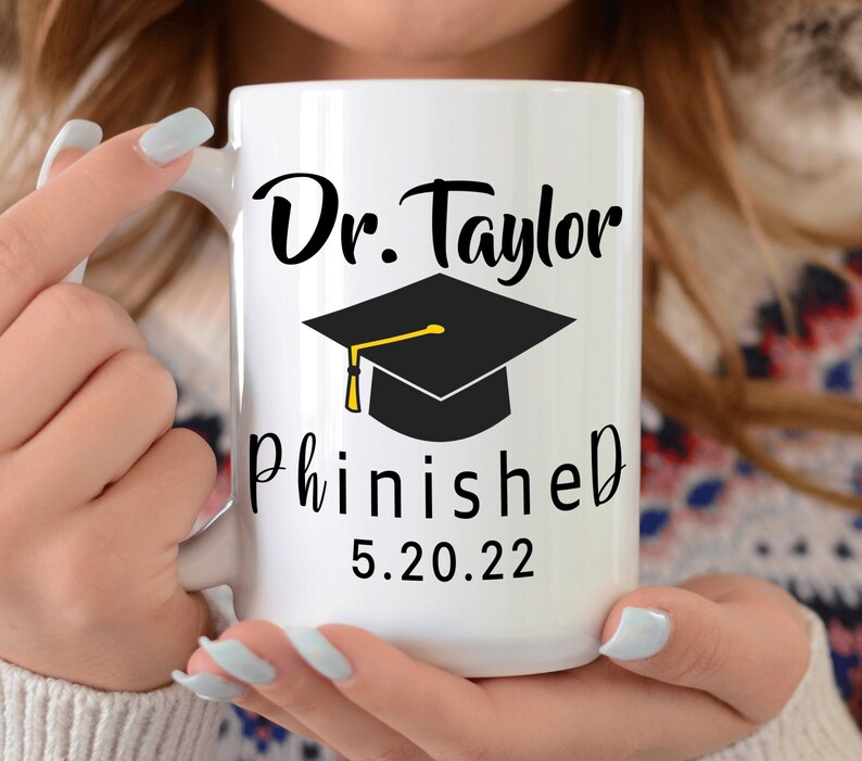 May include: White ceramic mug with a black graduation cap and the text "Dr. Taylor Phinished 5.20.22" in black.