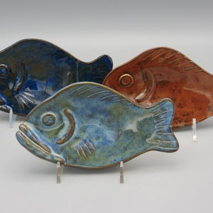 May include: Three ceramic fish-shaped plates in shades of blue and rust-orange. The plates have a glossy finish and are displayed on clear stands. The fish plates are a unique addition to any kitchen or dining room.
