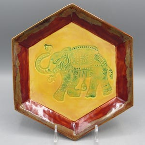 May include: Hexagonal ceramic plate with a yellow center featuring a green elephant design. The plate has a dark red border and a brown rim. The elephant is decorated with intricate patterns.