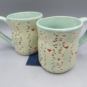 May include: Two ceramic mugs with a light green glaze. The mugs feature a birch tree design with black vertical lines and small red bird accents. Each mug has a curved handle and a light blue rim.