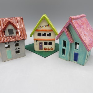 May include: Three ceramic house figurines. The left house has a red roof and gray walls. The center house features a green roof, orange trim, and painted pumpkins. The right house has a textured pink roof and light blue walls.