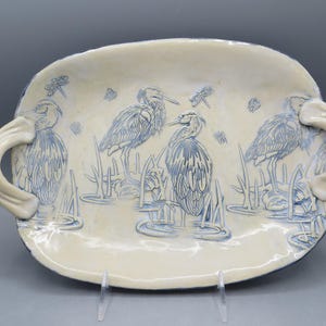 May include: An oval ceramic serving platter with a cream-colored glaze and blue embossed illustrations of three herons standing in water among reeds and dragonflies. The platter features two large, curved handles on the sides.