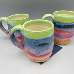 May include: Three ceramic mugs with colorful, layered glazes. The mugs feature a rainbow of colors, including green, blue, red, and yellow. Each mug has a handle and a light-colored interior. The mugs are handmade and suitable for hot or cold beverages.