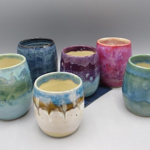 May include: A collection of six ceramic tumblers with rounded shapes and smooth, glazed finishes. The cups feature various color combinations, including shades of blue, green, purple, pink, and white, with some displaying drip-style glaze patterns.
