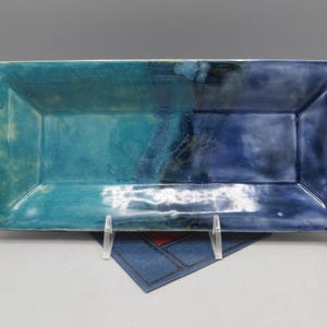 May include: A rectangular ceramic serving dish with a two-tone blue glaze. The left side is turquoise, and the right side is a darker blue. The dish has a slightly raised rim and is supported by a clear stand.
