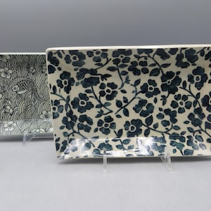 May include: Two rectangular ceramic serving platters. One features a pattern of fish and waves in black and white. The other has a cream background with a dark blue floral design. Both are displayed on clear stands.