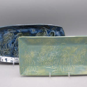 May include: Two ceramic serving platters. The larger platter is a deep blue with a heron design. The smaller platter is a light green with a heron design. Both platters have a rectangular shape and are displayed on clear stands.