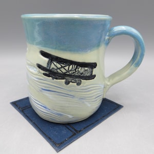 May include: Handmade ceramic mug with a light blue top and handle, featuring a black airplane design. The mug sits on a square blue coaster. The mug has a cream-colored base with blue wave-like accents.