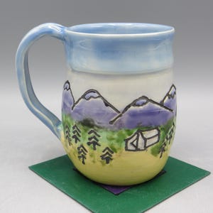 May include: A ceramic mug with a light blue handle and rim, featuring a hand-painted mountain and camping scene. The design includes purple mountains, green trees, and a white tent against a yellow and white background.