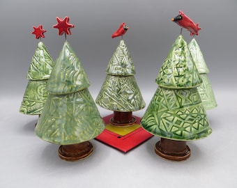 May include: A collection of ceramic Christmas trees in varying shades of green, adorned with intricate geometric patterns. Some trees are topped with red star ornaments, while others feature red cardinal bird figurines. The trees sit on brown bases.