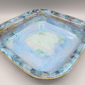May include: A square ceramic bowl with a glossy finish, featuring a blend of light blue, turquoise, and deep navy glazes. The rim is textured with mottled brown and dark blue tones, creating a distinct contrast against the smooth, pale blue center.