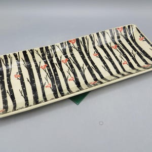 May include: A rectangular ceramic serving tray with a cream-colored base featuring a repeating pattern of vertical black tree trunks and small orange birds perched on the branches, finished with a glossy glaze and raised edges.