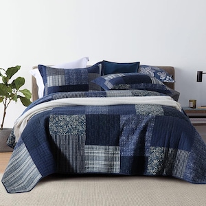 May include: A patchwork quilt featuring various shades of blue, including navy and light blue, with diverse patterns such as stripes, florals, and geometric designs. The quilt is draped over a bed with matching pillow shams and a white throw blanket.