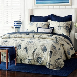 May include: A white and blue nautical themed quilt with a seashell and starfish pattern. The quilt is on a bed with blue and white pillows and a white headboard. The bed is in a bedroom with a white bedside table and a blue rug.