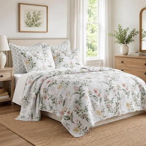 May include: A white quilt and matching pillow shams featuring a botanical print with green leaves and soft pink and yellow flowers. The bedding set has a quilted texture and is draped over a bed with a beige upholstered headboard.