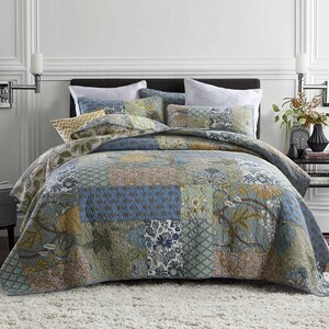 May include: A patchwork quilt and pillow set in blue, green, and tan. The quilt has floral and geometric patterns. The bed is made up with white pillows and a dark headboard. The quilt is on a white rug.