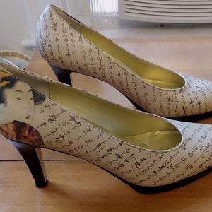 Puede incluir: Un par de zapatos de tacón alto con fondo blanco y escritura negra. Un zapato tiene un diseño inspirado en la geisha con un patrón floral rojo y dorado.