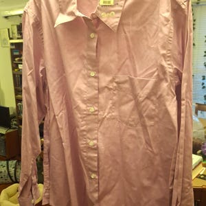 May include: A light pink button-down shirt with long sleeves, a chest pocket, and a collar. The shirt is hanging on a hanger. A tag is attached to the collar. The shirt is made of a smooth fabric.
