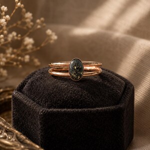 May include: A rose gold ring featuring a double band design and a central oval-shaped setting. The oval stone is dark green with embedded gold-colored flecks, creating a speckled appearance within the smooth, polished metal frame.