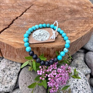 Peut inclure: Un bracelet de perles turquoise et noires avec une étiquette « Suellarts ». Le bracelet est présenté sur une surface en bois avec un fond de roches et de fleurs. Le bracelet est composé de perles de turquoise et de lave noire.