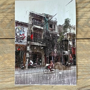 Könnte beinhalten: Ein Vintage-Foto einer Straßenszene in Hanoi, Vietnam. Das Bild ist verblasst und zerkratzt, was ihm einen abgenutzten Look verleiht. Das Foto zeigt ein Gebäude mit einem Schild, auf dem "Hanoi City Palace" steht. Es sind Menschen und Fahrräder auf der Straße zu sehen.