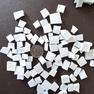 May include: A collection of small, white, square and rectangular mosaic tiles scattered on a brown surface. A U.S. quarter coin is included for size reference. Some tiles are in a clear plastic bag in the background. Ideal for craft projects.