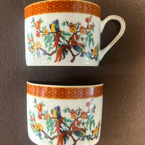 May include: A white teacup and matching saucer with a decorative bird and floral design. The cup has a gold handle and rim. The rim of both pieces has a geometric orange pattern. The design features colorful birds, flowers, and branches.