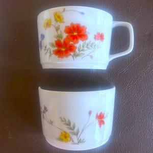 May include: Two white ceramic mugs with floral designs. One mug has a handle. The floral pattern includes orange, yellow, and red flowers with green leaves. The mugs are set against a brown background.