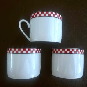 May include: Three white ceramic cups with a red and white checkered pattern around the rim. One cup has a heart-shaped handle. The cups are arranged on a dark brown surface.