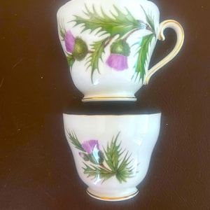 May include: Two white teacups with a thistle design. The cups feature a gold rim and handle, and the thistle design includes purple flowers and green leaves. The cups are placed on a brown surface.