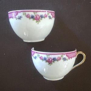 May include: Two pieces of a white teacup with a pink floral design. The rim of the cup has a pink lace pattern. The cup has a gold handle. The teacup is broken.