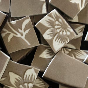 May include: A close-up of a pile of brown ceramic tiles with a white floral design. The tiles are broken into irregular shapes and sizes.