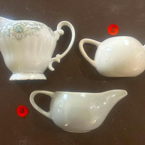 May include: Three white ceramic serving pieces: a creamer with floral accents, a small teapot, and a gravy boat. The creamer has a decorative handle and a scalloped base. The teapot is round with a curved spout. The gravy boat has a curved handle.